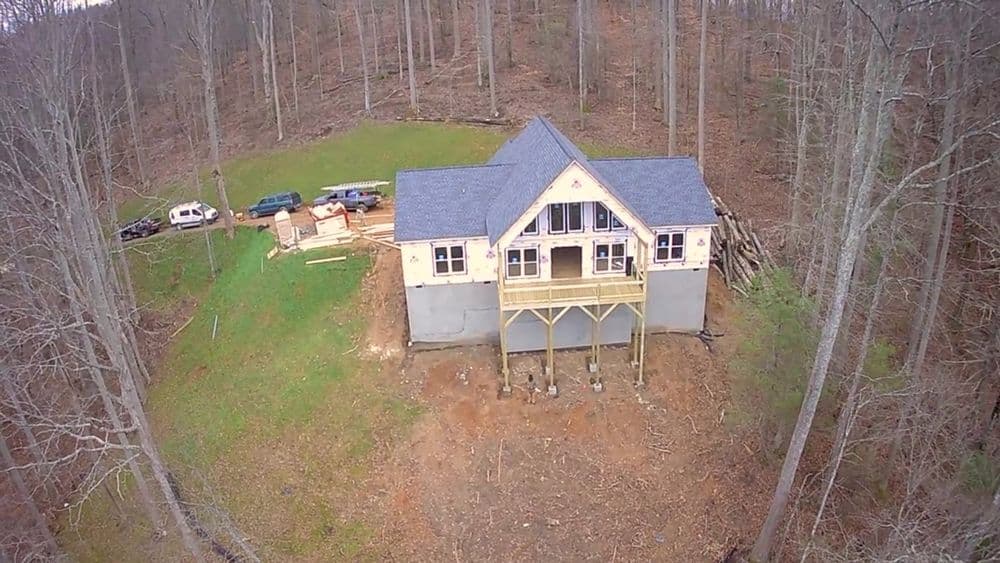 Aerial view of a modern house under construction in a wooded area with vehicles nearby.