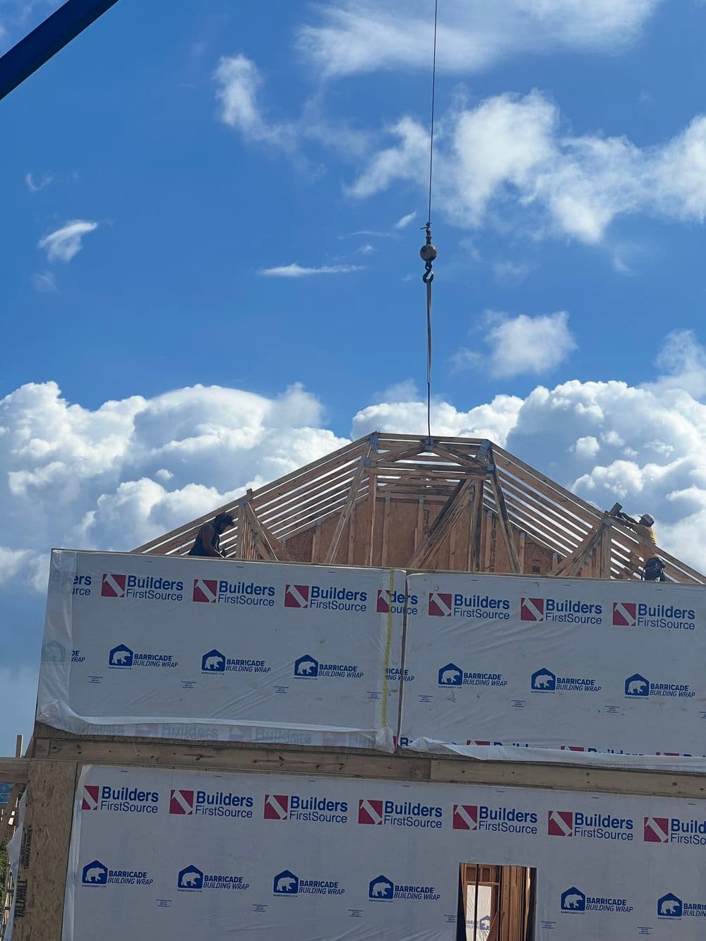 Construction site with wooden roof frame being lifted, surrounded by clouds.