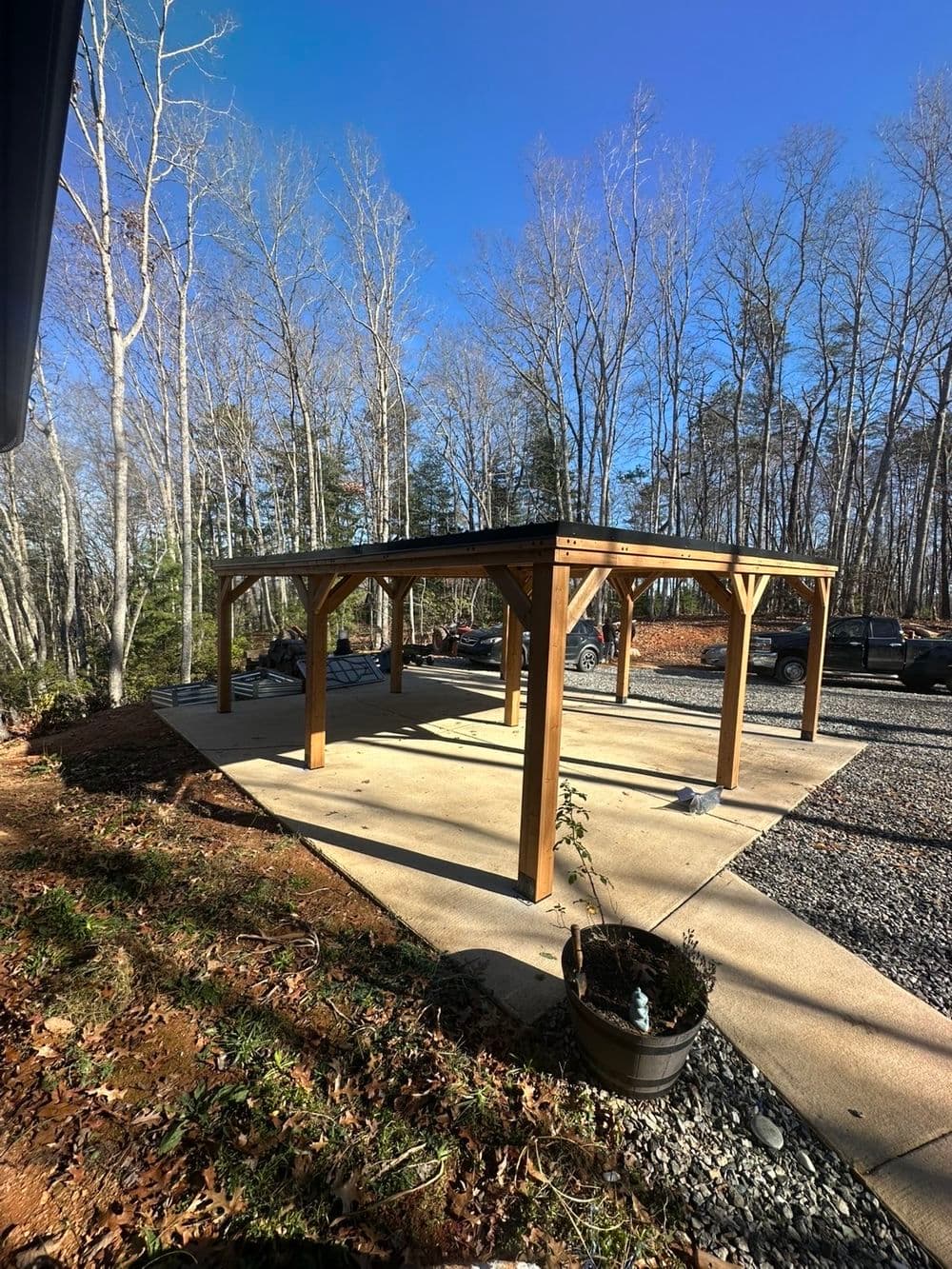 Wooden carport structure in a wooded area, with gravel driveway and parked vehicles nearby.