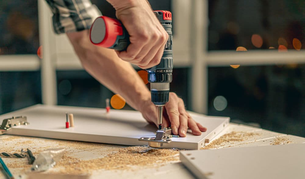 Person using a cordless drill to assemble furniture on a worktable with wood shavings.